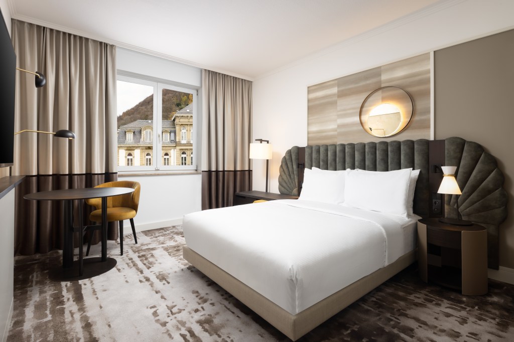 Hilton Heidelberg Queen Deluxe Room, Queen bed with unique shaped headboard in a room with light fixture above and next to the bed and a yellow chair with a table and a window looking out to the city