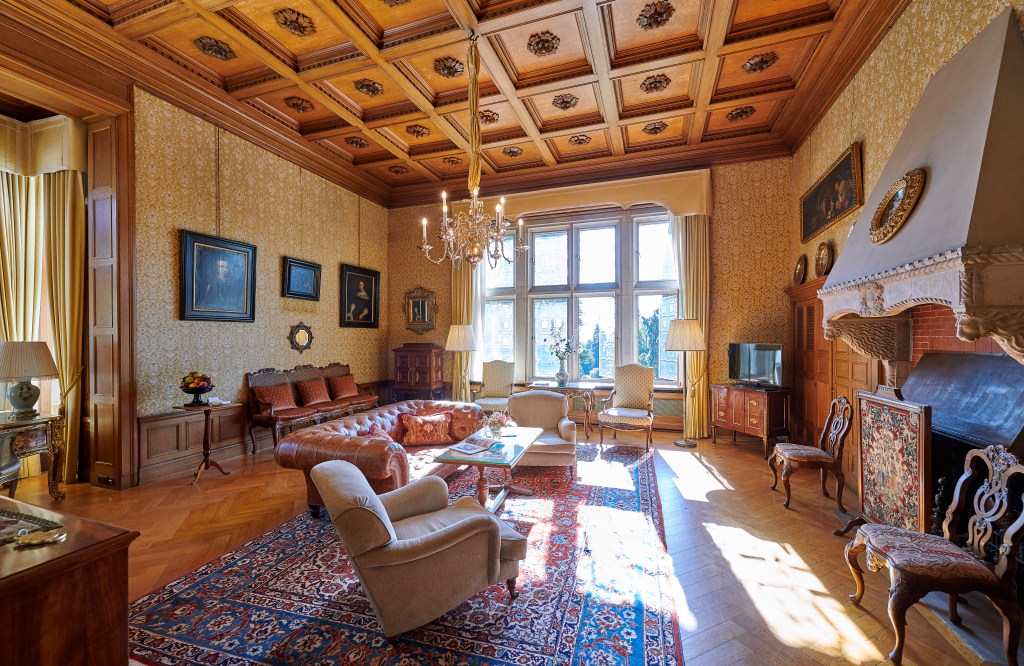 Schlosshotel Kronberg, an SLH Hotel Royal Suite Living Room, Suite living room with lounge seating on an area rug in a room with wooden crown molding on the ceiling and a chandelier hanging as well as framed portraits on the wall and a fireplace with a grand mantel
