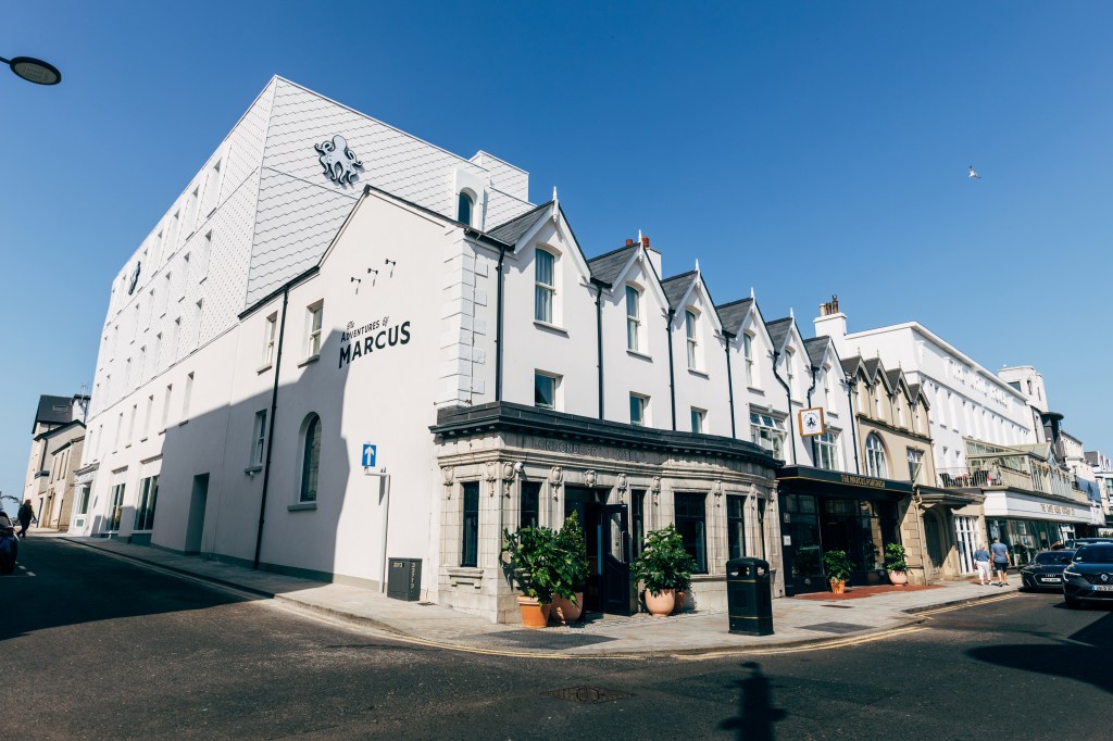 Exterior of The Marcus Portrush, Tapestry Collection by Hilton - white multi-story building on a street corner with "The Adventures of Marcus" written on the side of the building