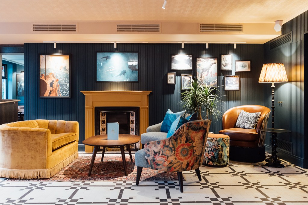 Lobby with yellow sofa, colorful patterned chairs, fireplace, black and white tile floor, nautical-themed artwork on a navy wall at The Marcus Portrush, Tapestry Collection by Hilton - Lobby