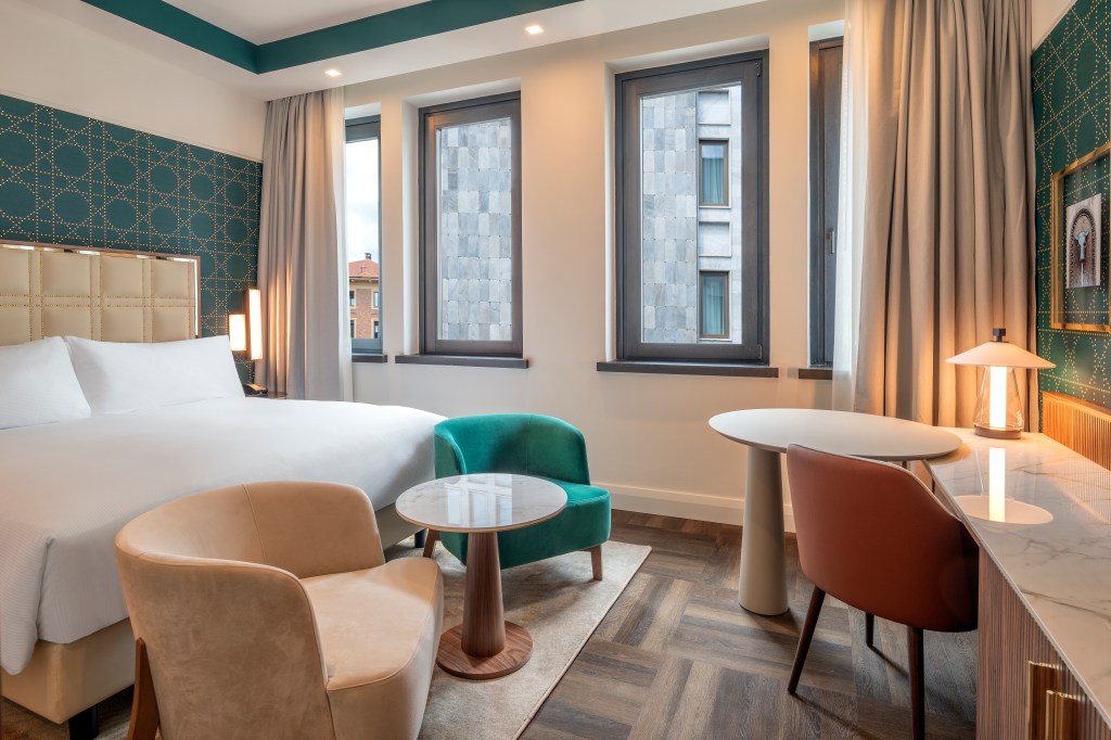 Hilton Turin Centre King Guest Room Bedroom, bed, two chairs and middle table, table and chair desk area windows with view of hotel and city