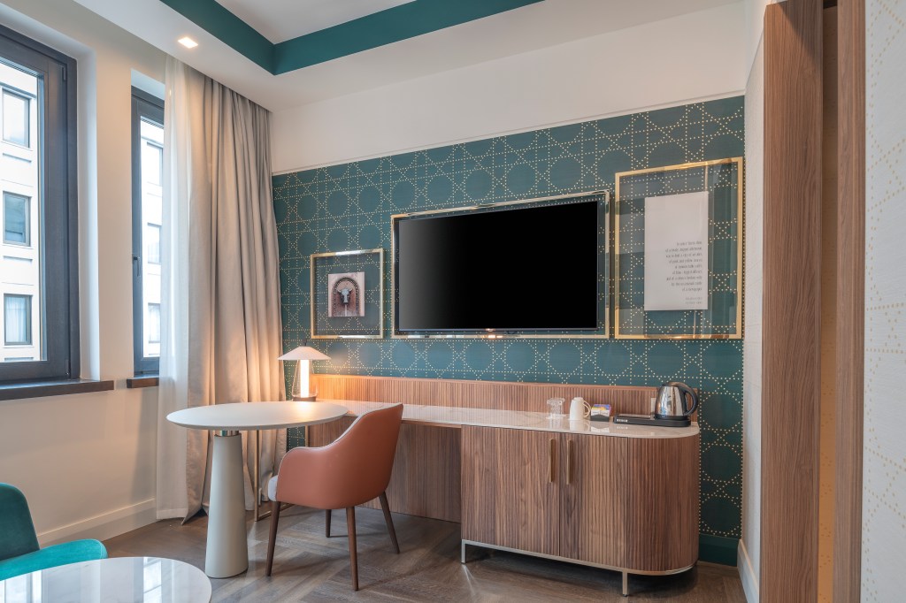 Hilton Turin Centre King Guest Room Bedroom, table and chair facing windows with small lantern, tea kettle on entertainment stand below tv and decorative framed images
