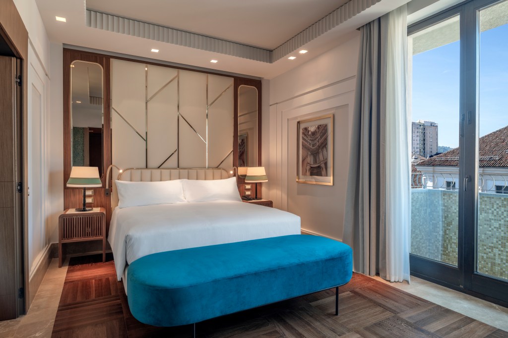 Hilton Turin Centre Presidential Suite Bedroom, side tables with lamps, bed with white linens and blue bench at the end, balcony with view of city