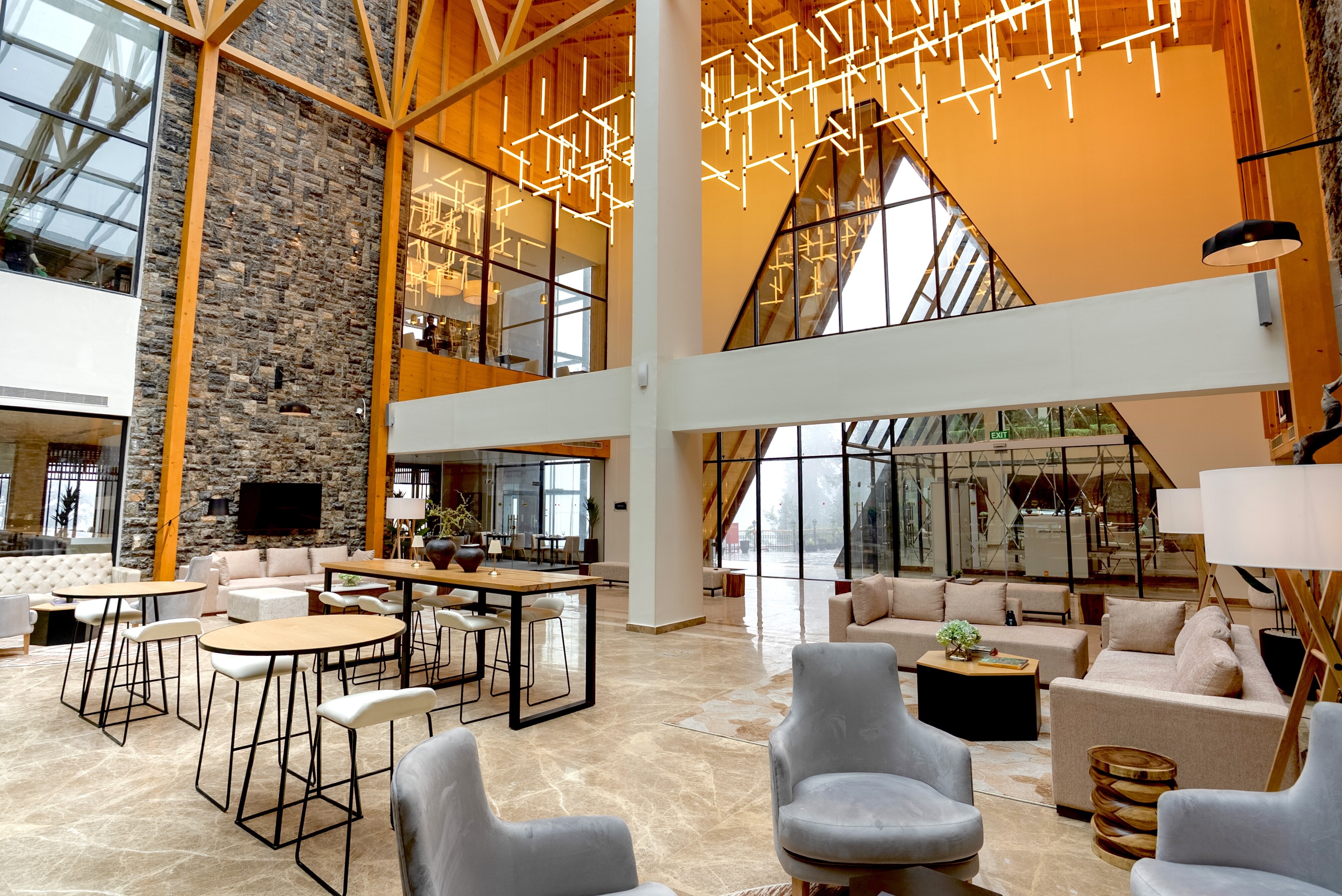 DoubleTree by Hilton Nathiagali Lobby with a lounge couch sitting areas and a lounge chair sitting areas and high top table sitting areas in a room with cathedral ceilings with modern light fixtures and an exposed stone wall next to orange post and beams and a a-frame shaped wall of windows