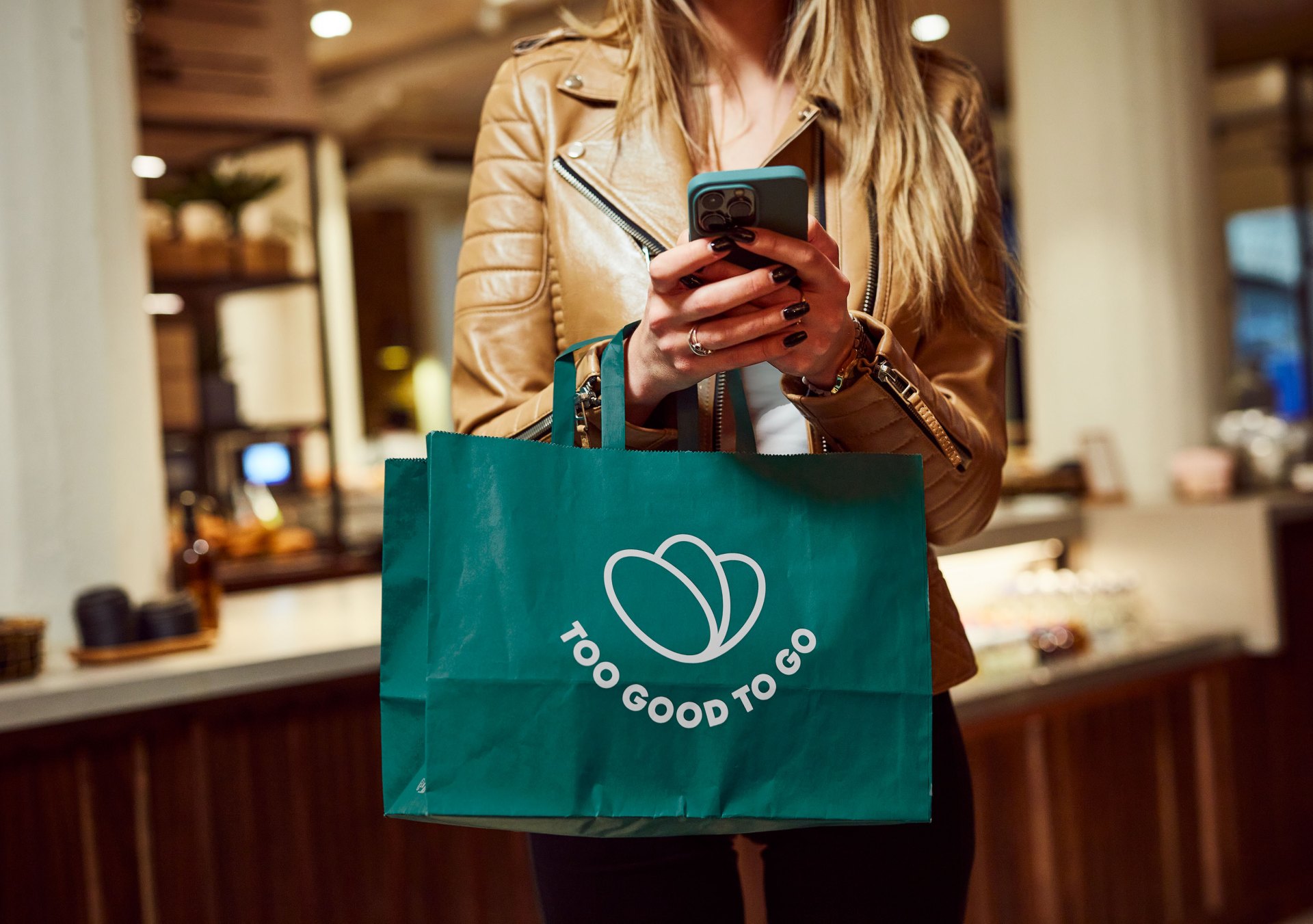 A woman holding a phone and a Too Good To Go bag