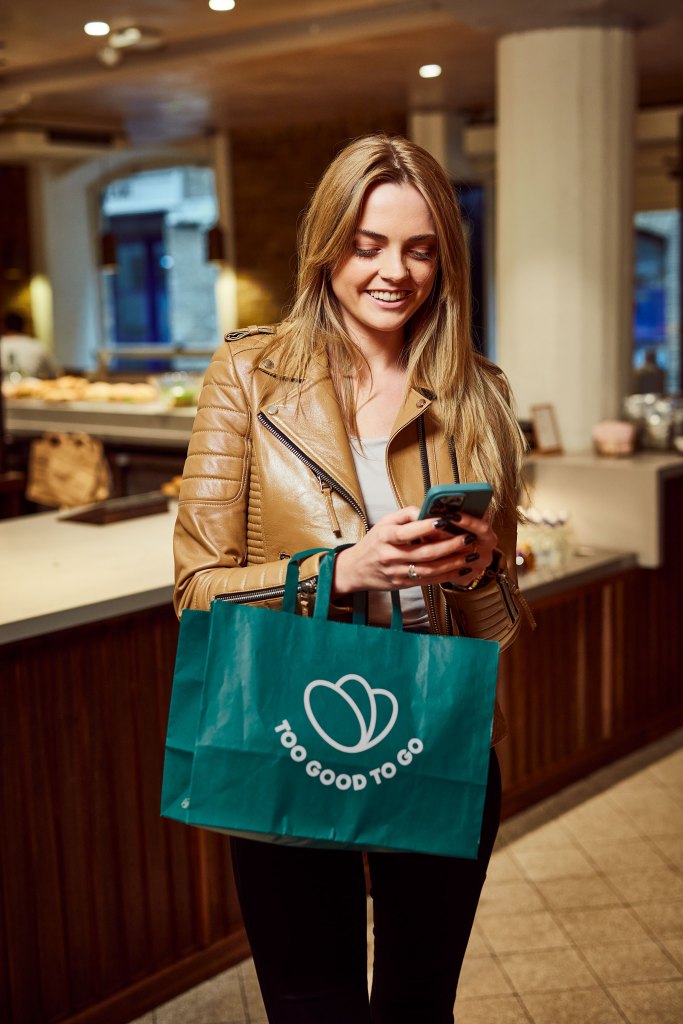 A woman holding a phone and a Too Good To Go bag