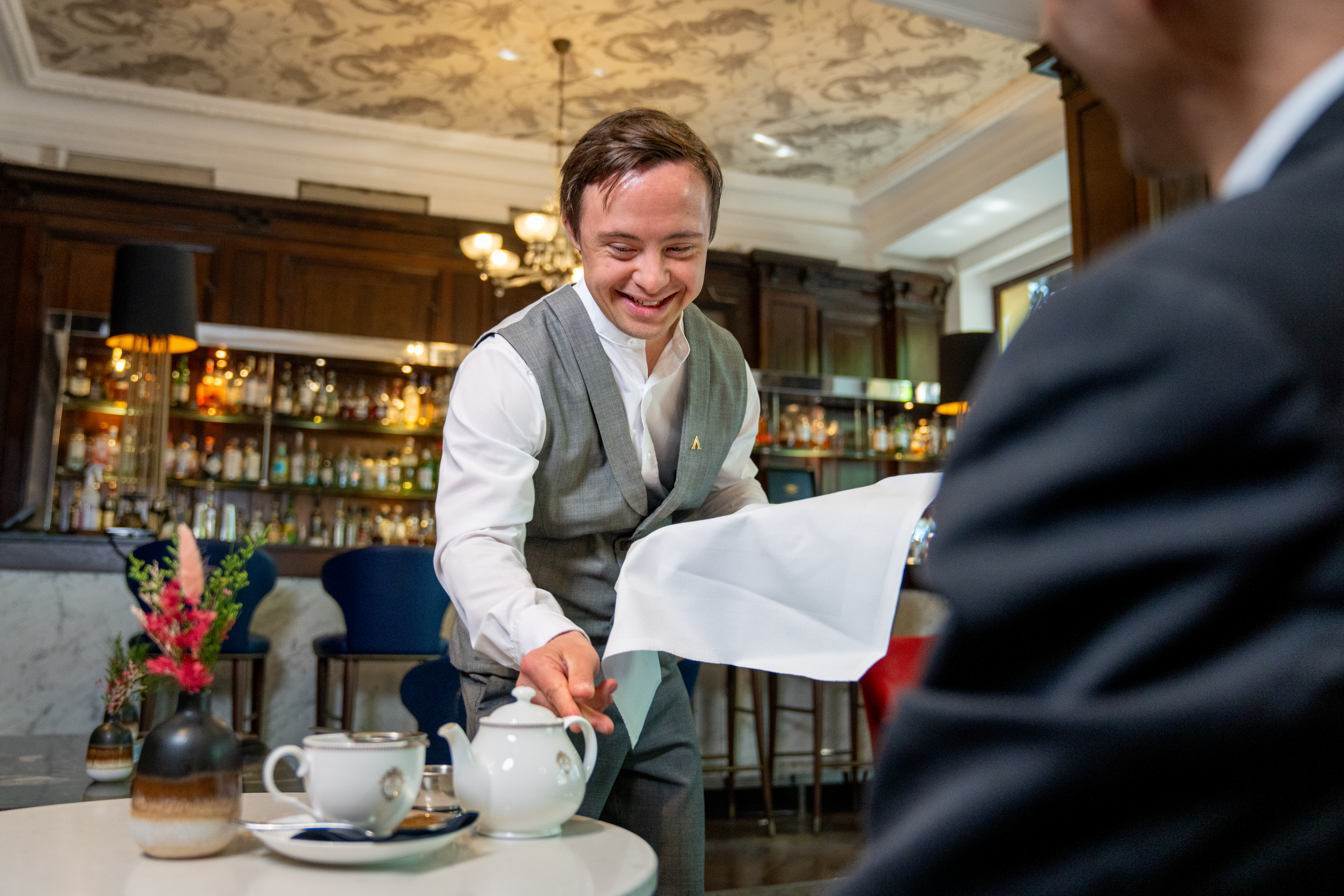 Alban placing a teapot on a table in front of a guest