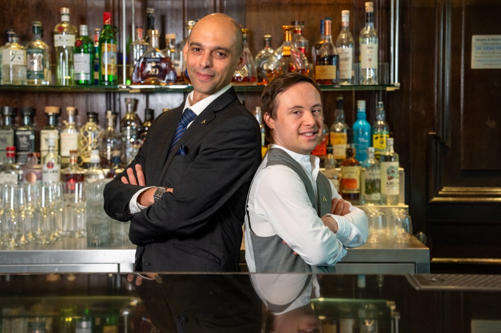 Alban and another man stand back to back behind a bar smiling at camera