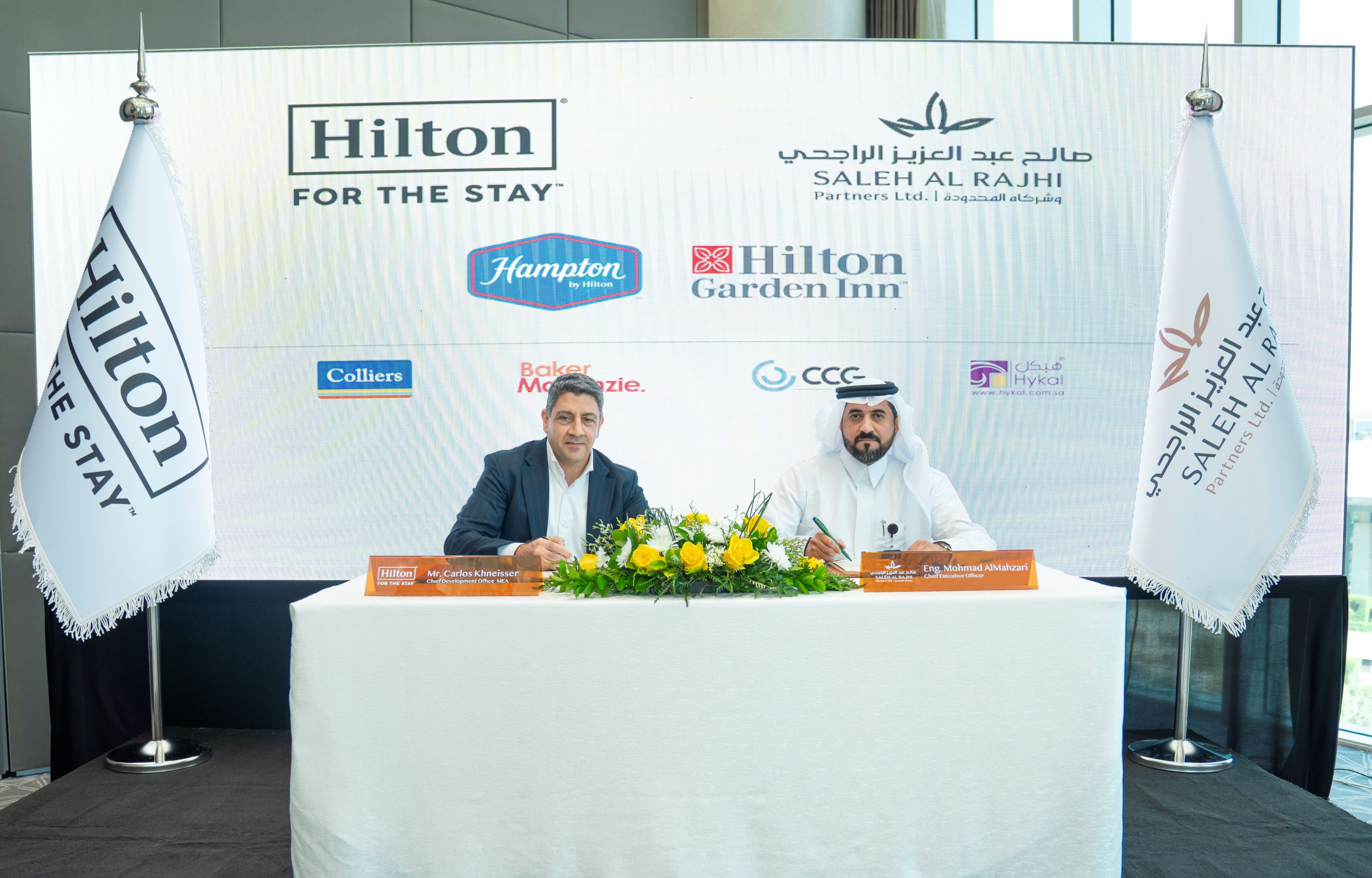 Carlos Khneisser and Mohmad AlMahzari set at a table signing paperwork in front of a Hilton background