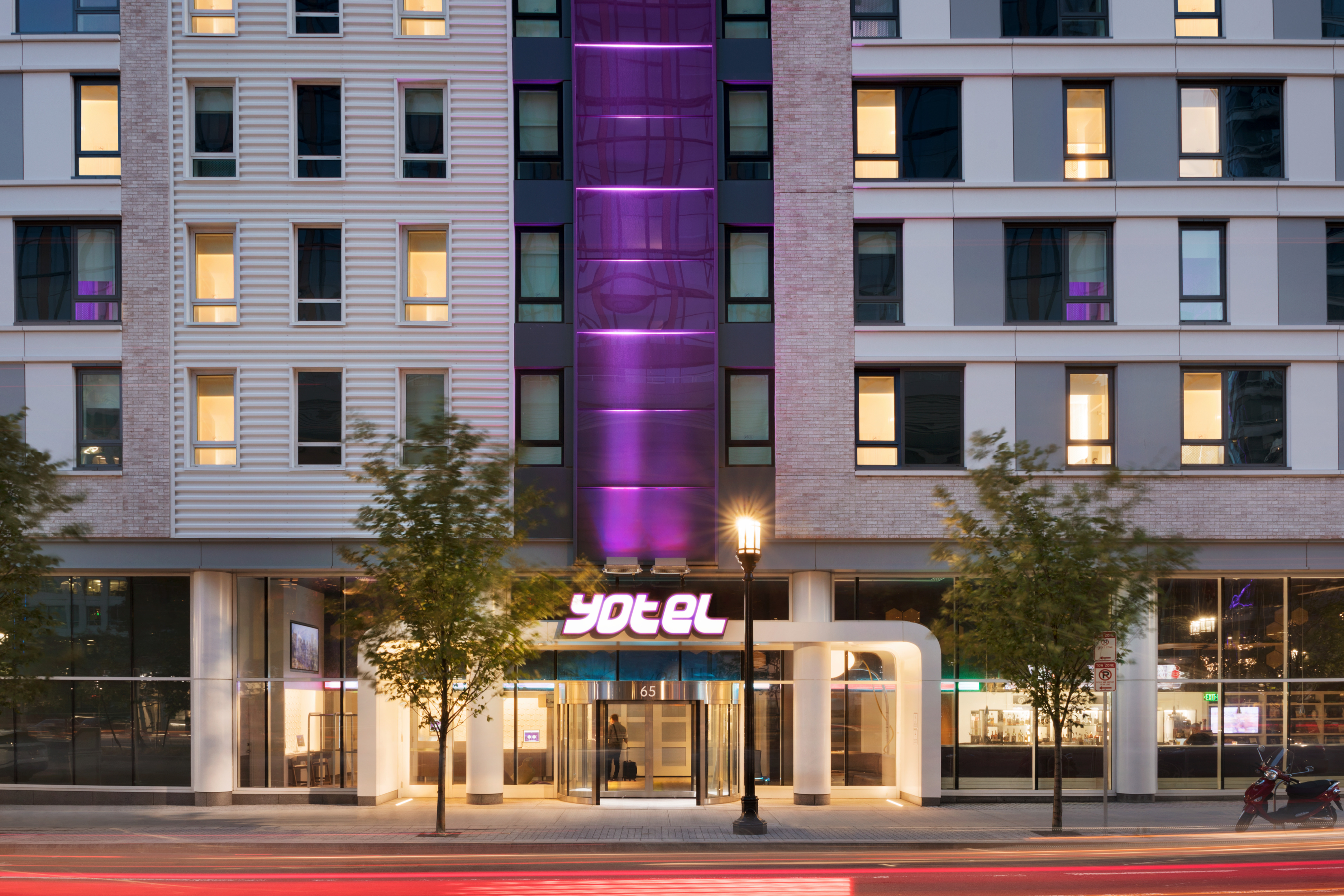 Modern hotel entrance with revolving glass doors, illuminated Yotel sign, streetlamp, trees, and light trails from passing traffic at night.