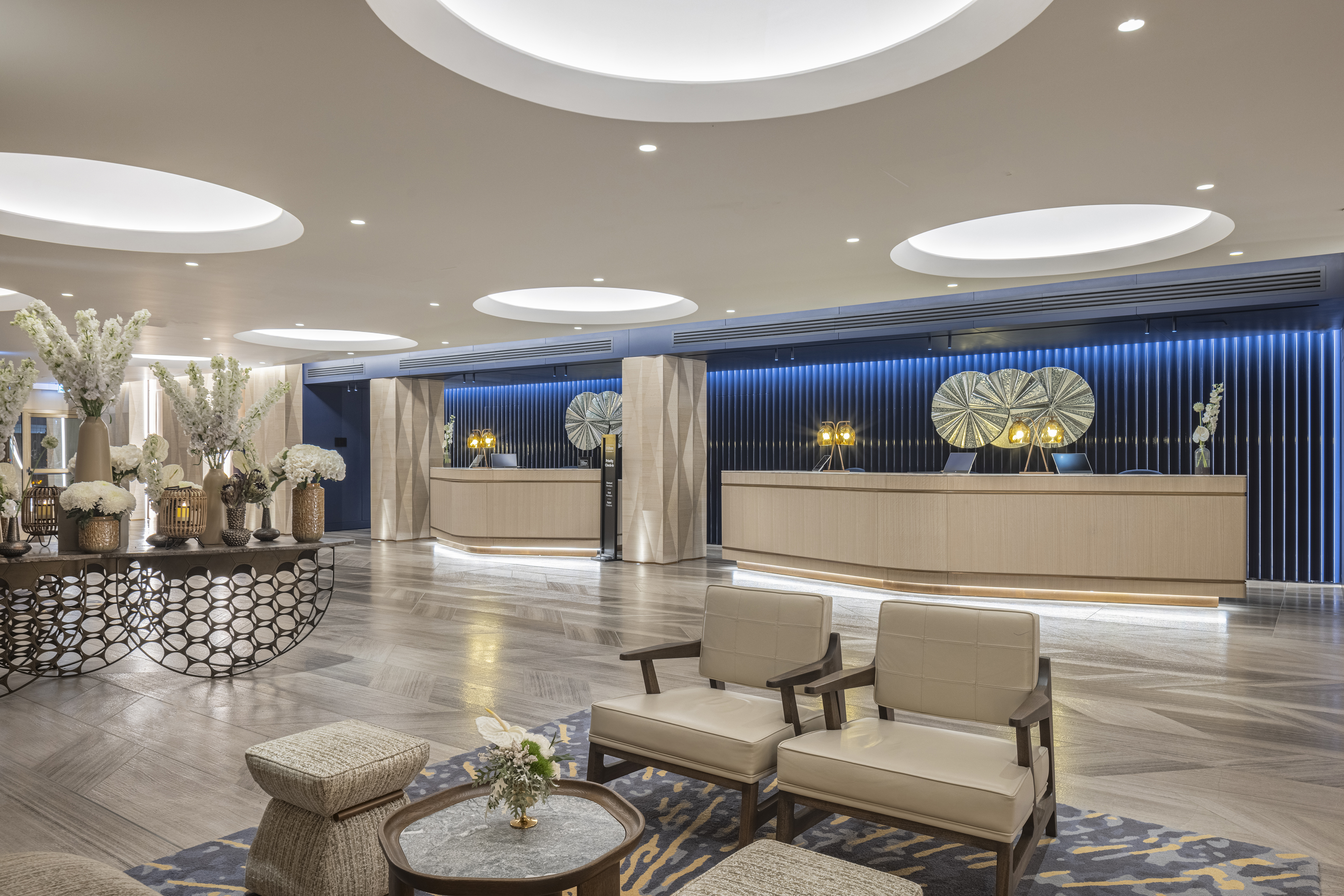 Modern hotel lobby with two beige reception desks, decorative lighting, floral arrangements, and seating area with chairs and a table.