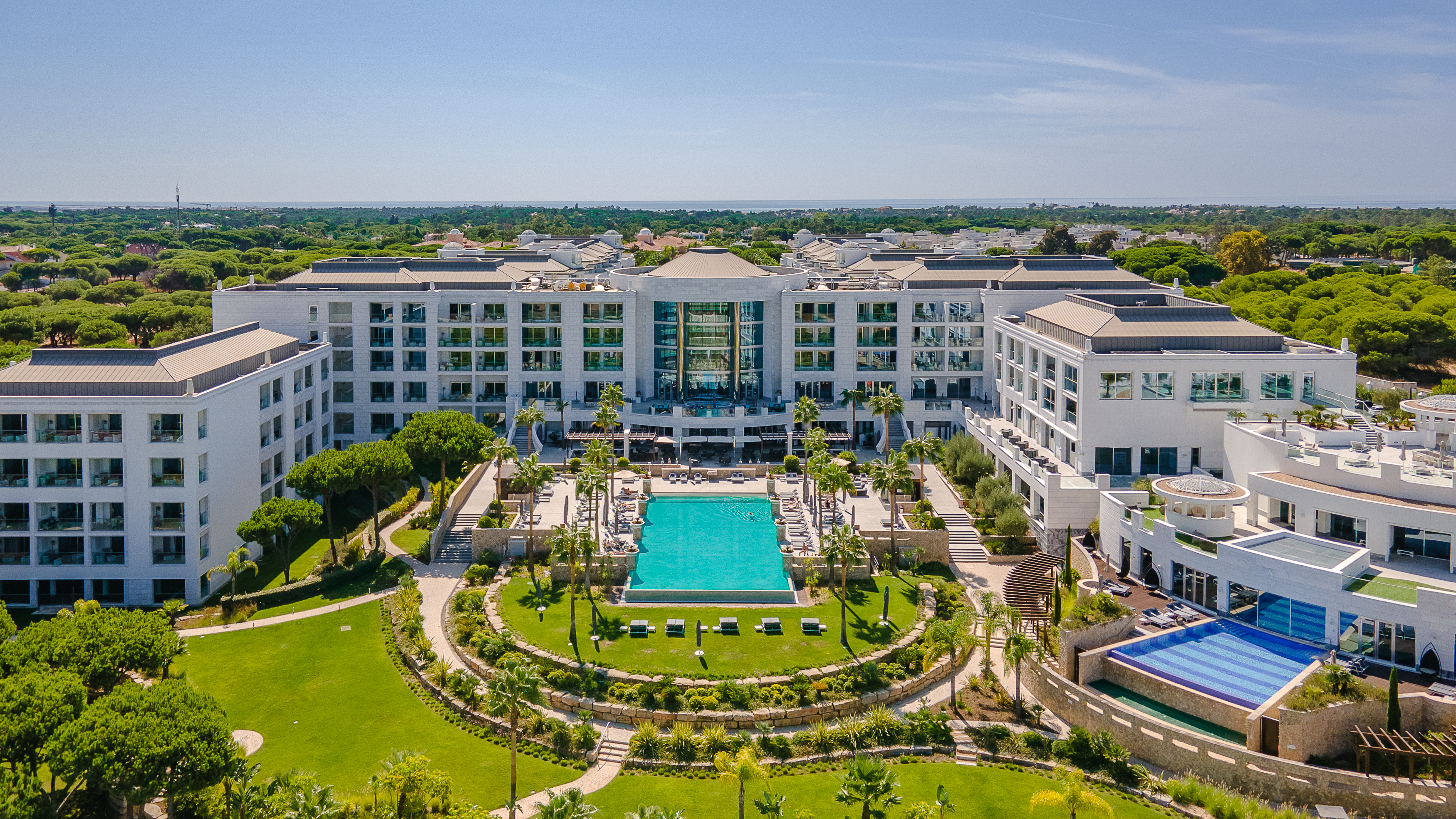 Luxury resort with a large central swimming pool, surrounded by palm trees and manicured gardens. White multi-story buildings frame the pool area.