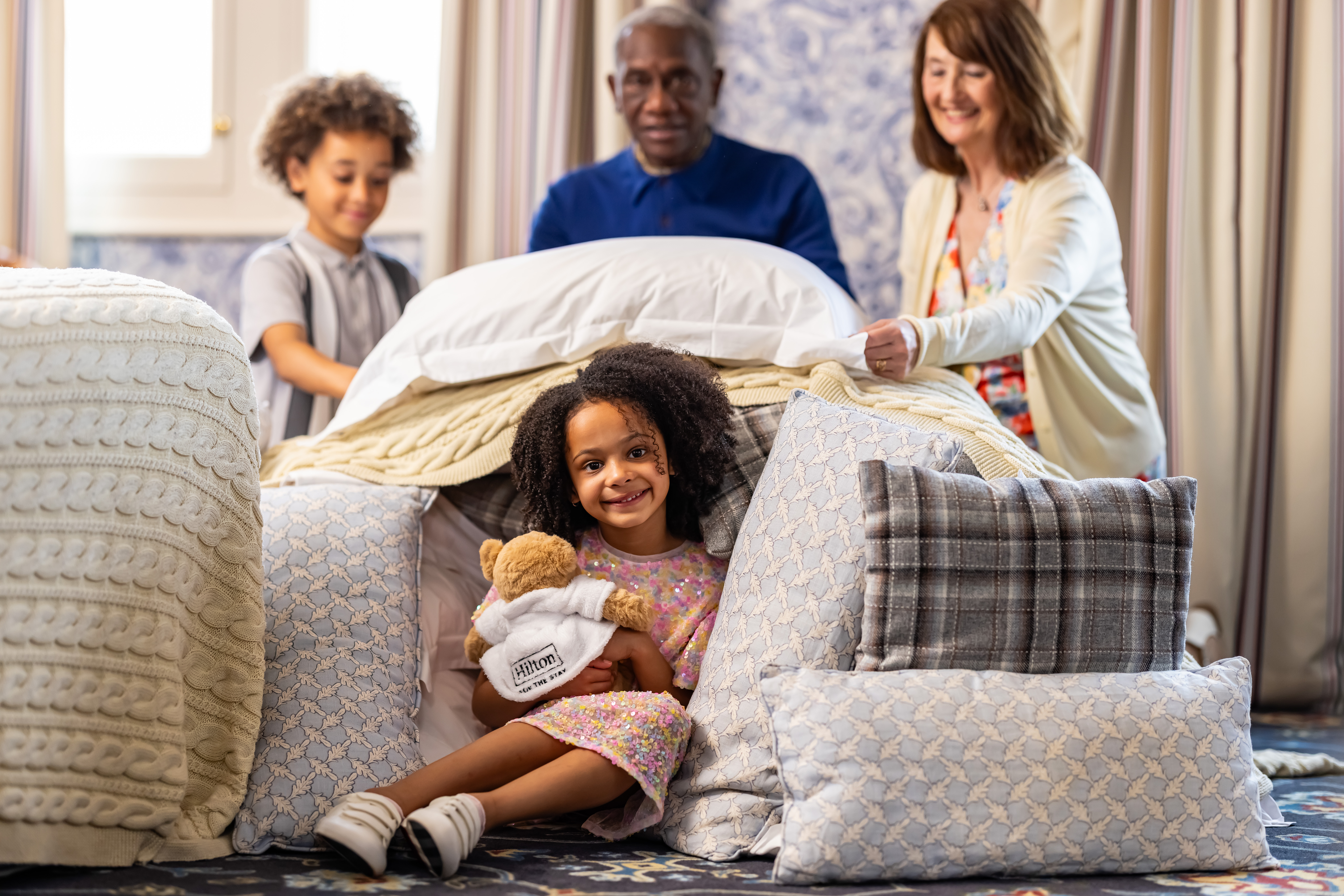 Young girl holding a teddy bear sitting inside a pillow fort, with two adults and another child smiling behind her.