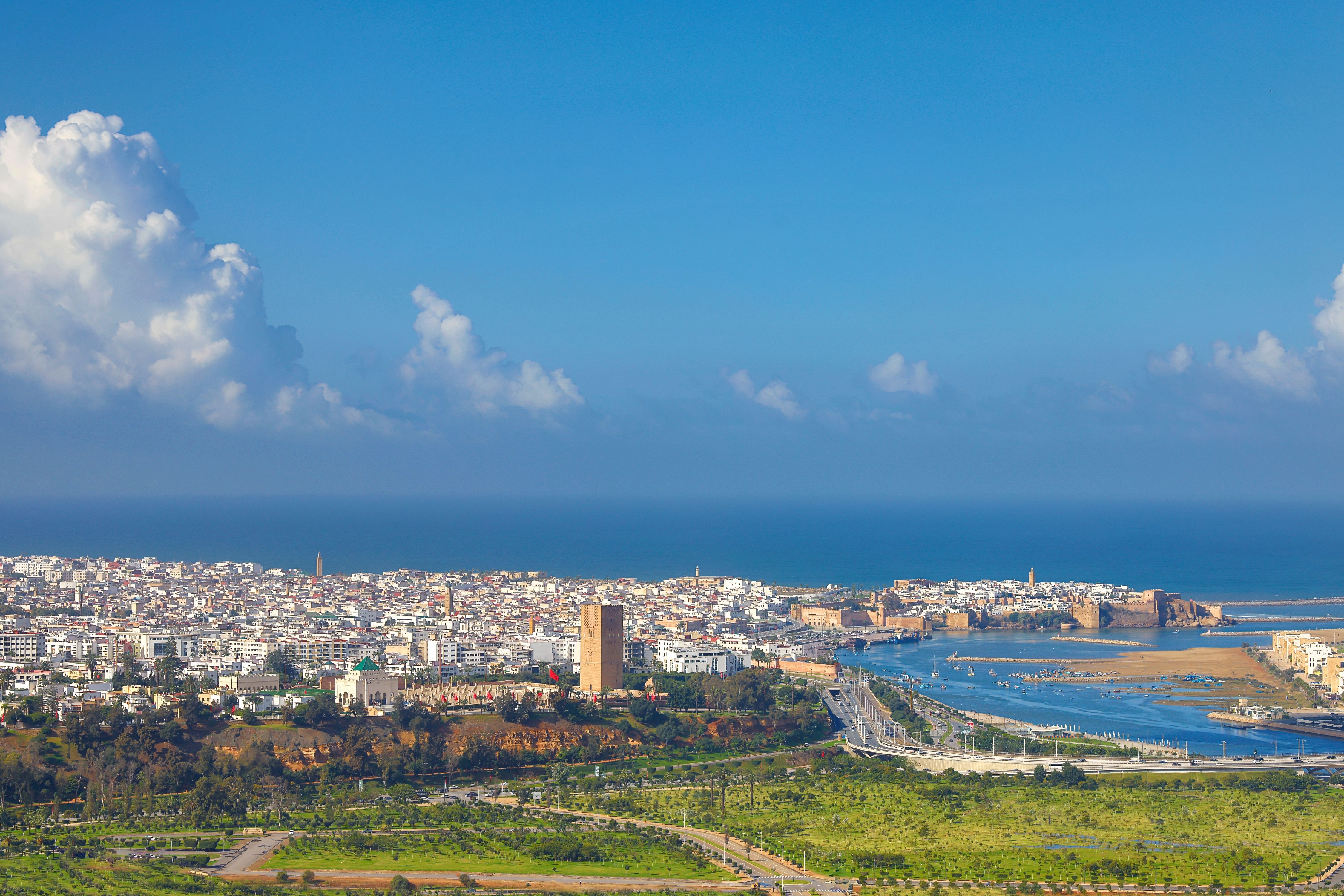 Coastal cityscape with a river, historic buildings, and a large tower under a blue sky with scattered clouds.