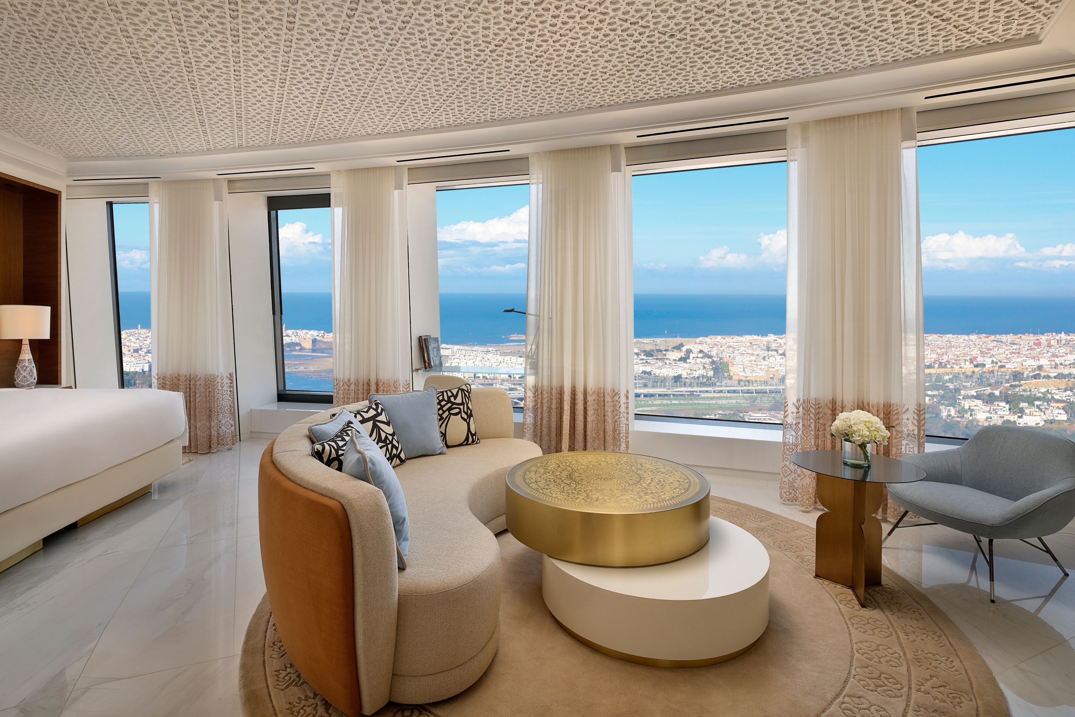 Luxurious living area with curved sofa, decorative coffee tables, large windows with ocean view, and patterned ceiling.