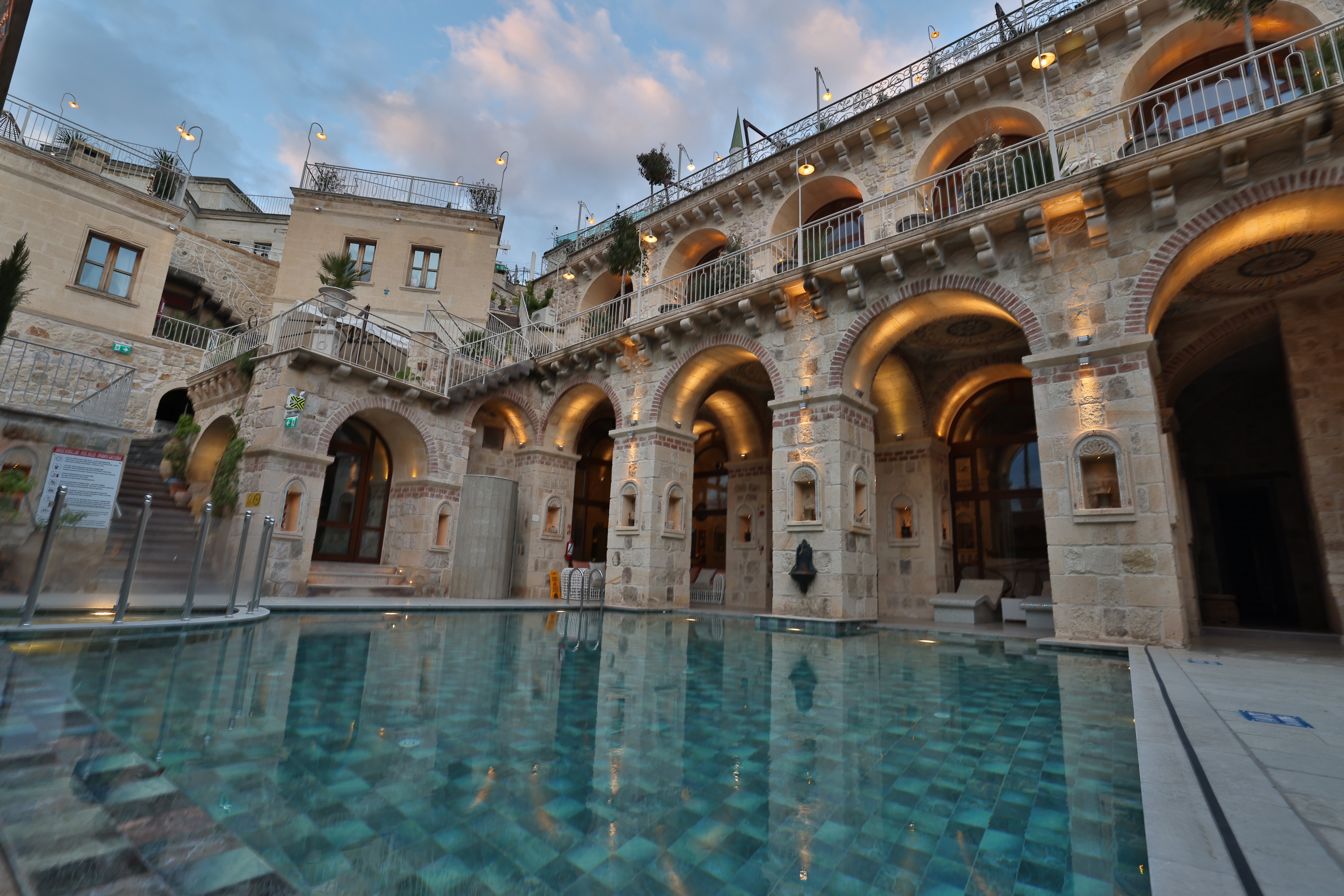Stone building with arched doorways and warm lighting surrounding a clear blue tiled swimming pool at dusk.