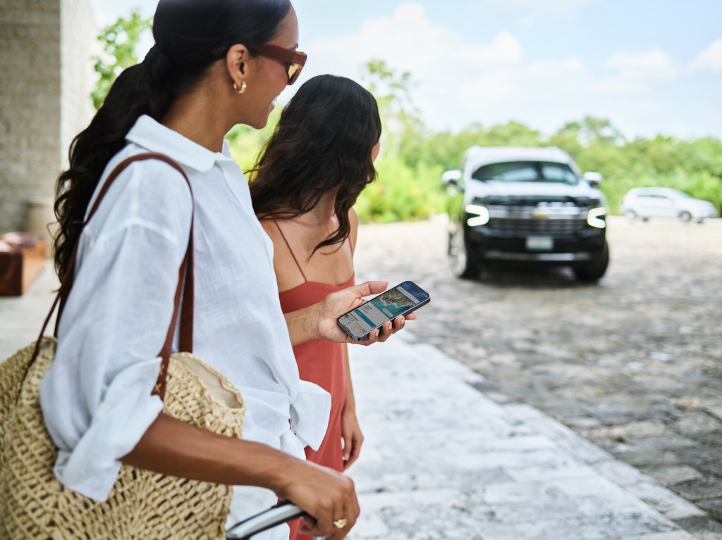 Two women looking at a smartphone while a black car approaches on a cobblestone driveway.
