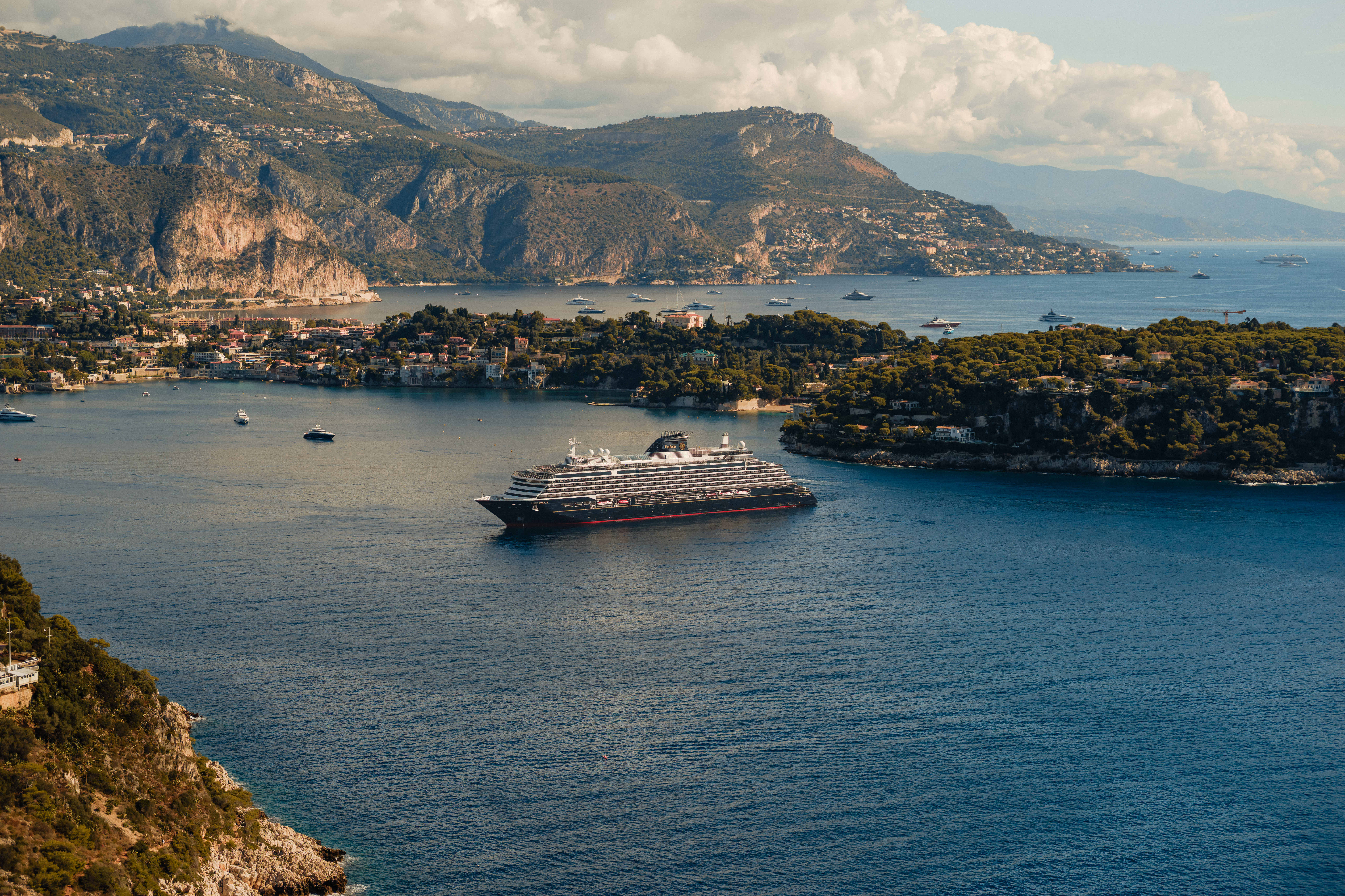 Explora Journeys EXPLORA II Cruise ship sailing in a coastal bay surrounded by green hills and rocky cliffs under a partly cloudy sky.
