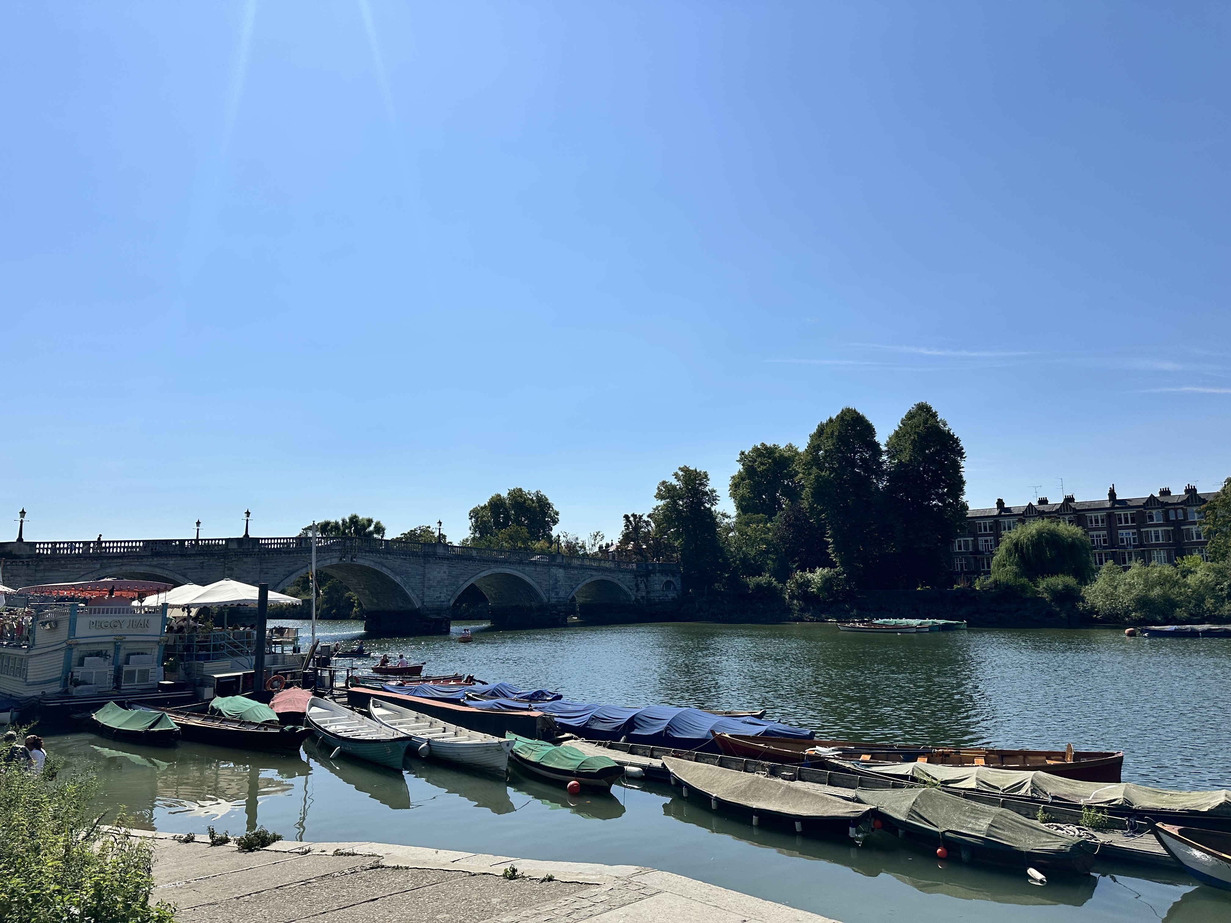 River with moored boats, a stone bridge in the background, and trees and buildings along the riverbank under a clear blue sky.