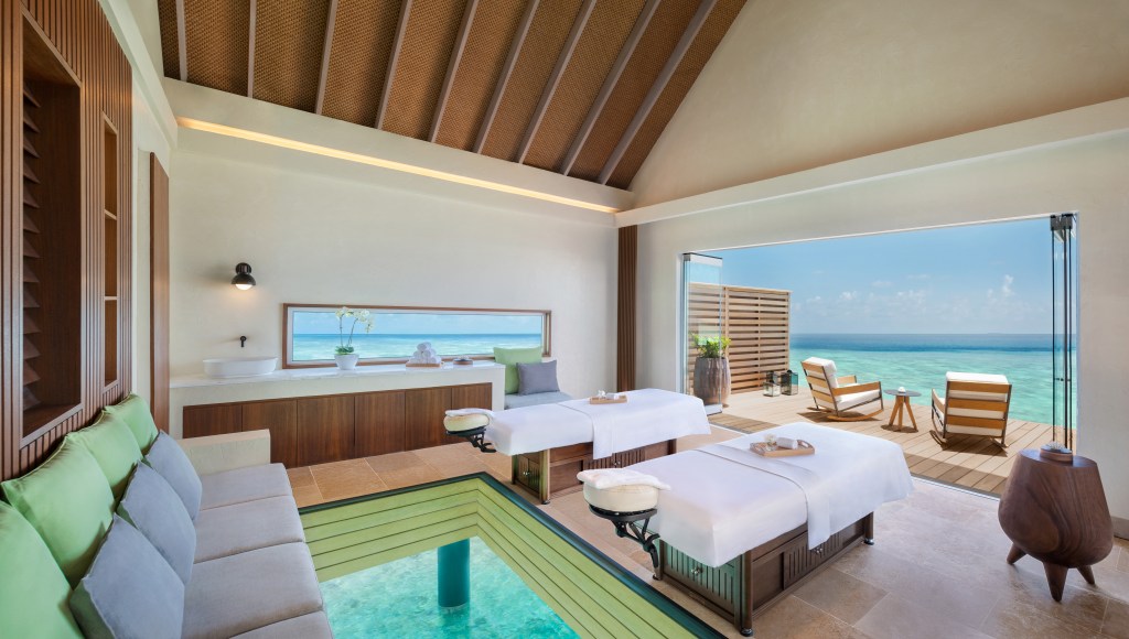 luxurious spa room a couch, sink area, two spa beds with outdoor deck with table and chairs overlooking the ocean