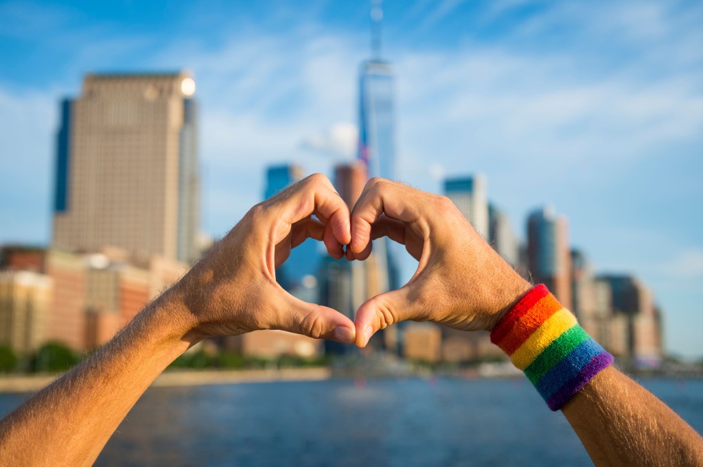 Hands making heart symbol in front of city skyline wearing Pride rainbow sweat band