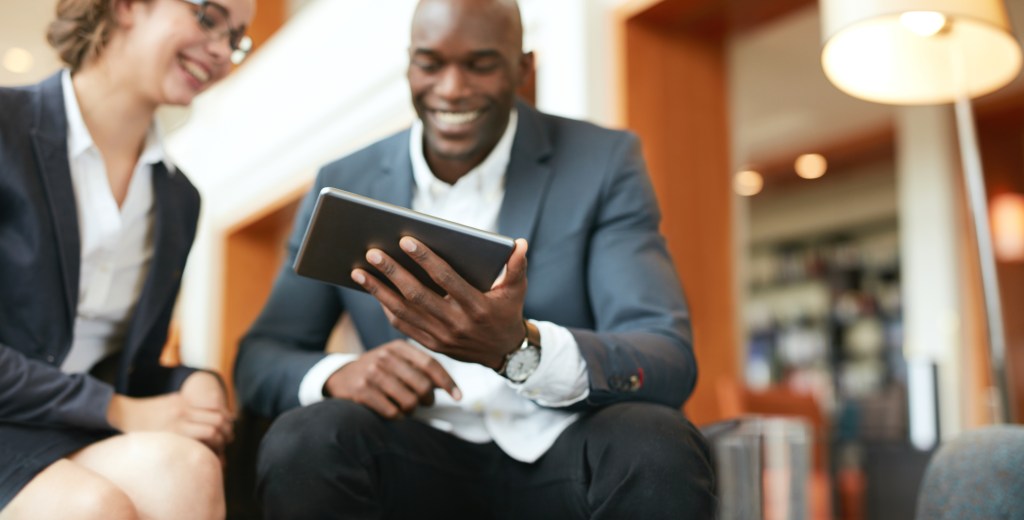 Happy young business people sitting together using digital tablet while at hotel lobby. Focus on tablet computer.