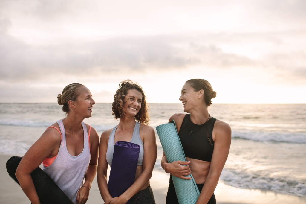 Smiling women at the beach talking to each other holding yoga mats