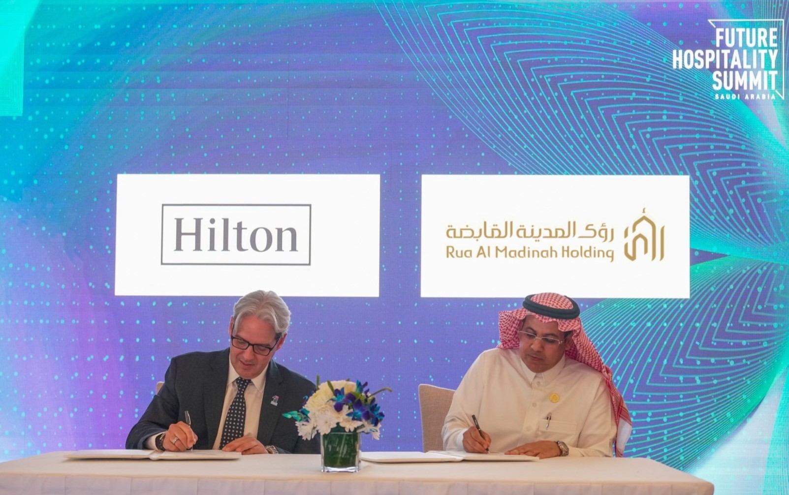 Rua Al Madinah Hilton Signing - two men sitting at a table, signing papers, decorative flowers in the center, Hilton and Rua Al Madinah Holding and Future Hospitality Summit Saudi Arabia logos in the background