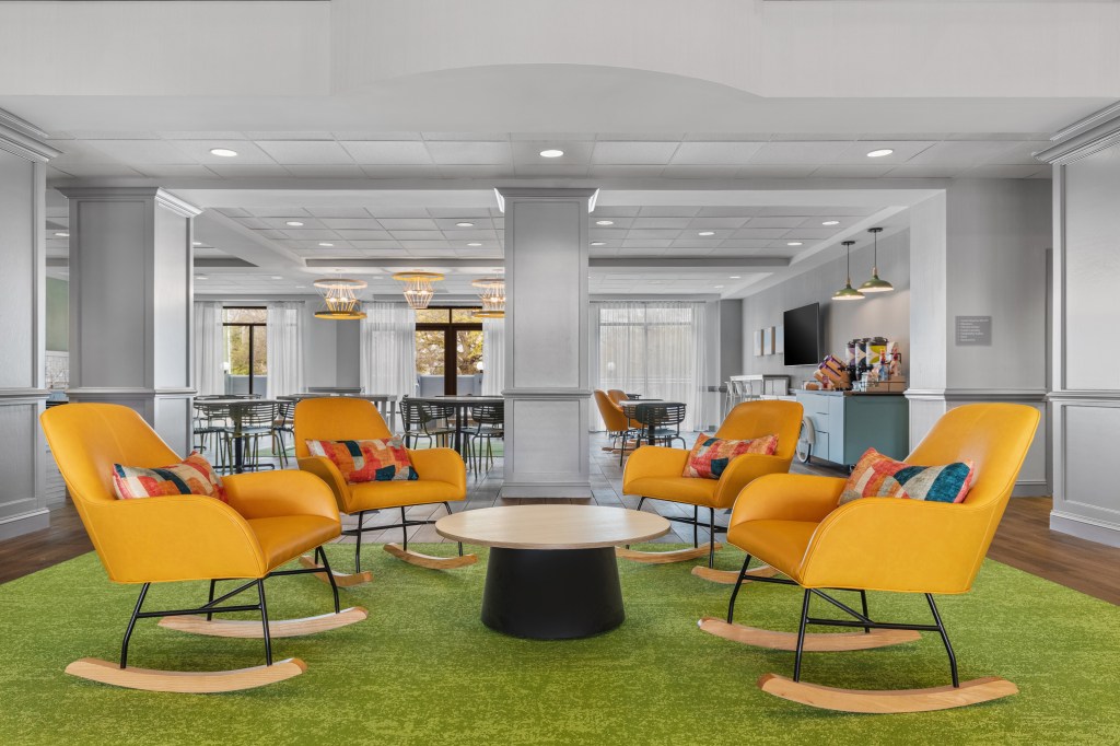 Yellow rocking chairs with colorful pillows around a table and green carpet in the lobby of Spark by Hilton Charlotte Tyvola I-77