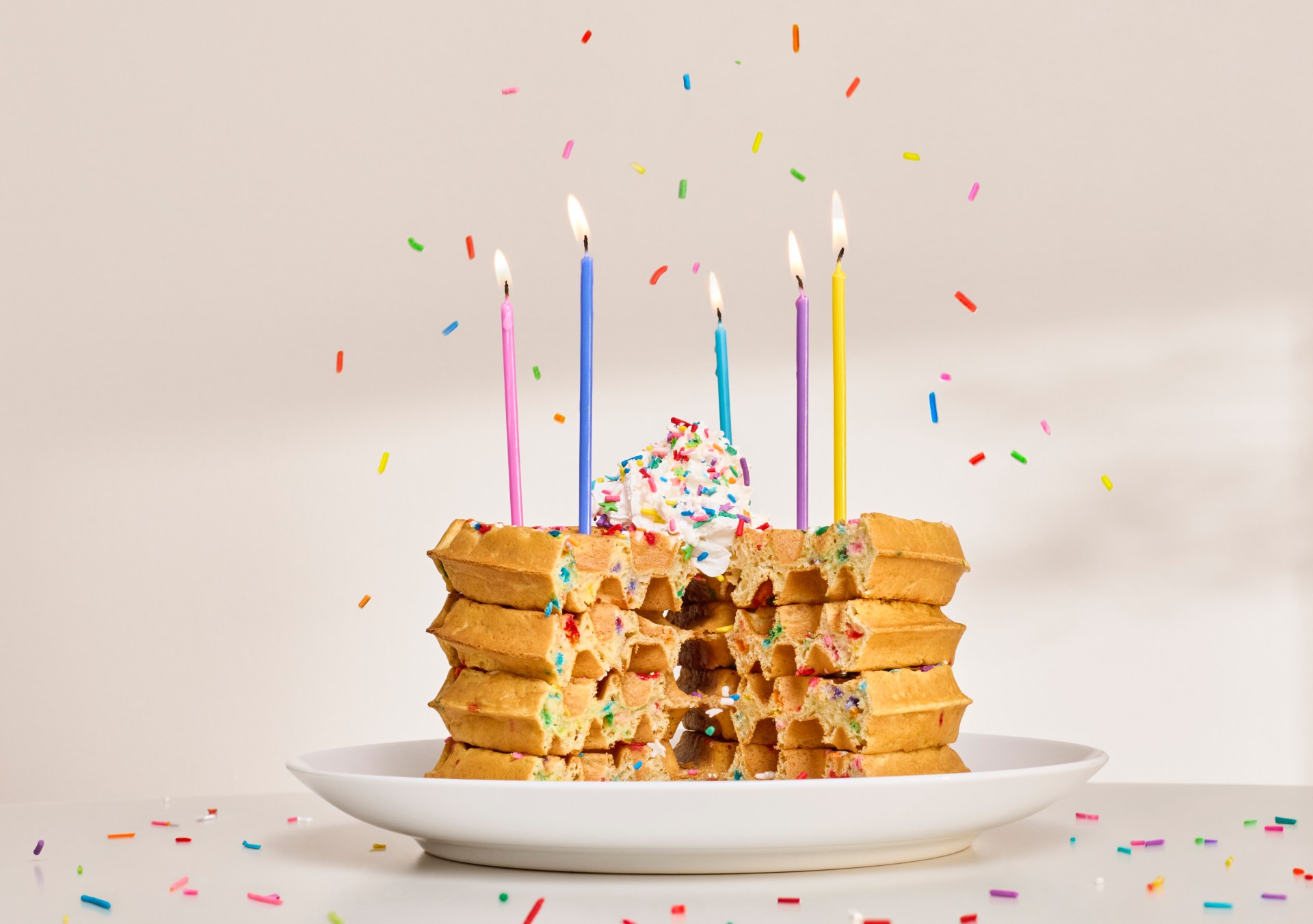 stack of 4 birthday cake flavored waffles with whipped cream, sprinkles and 5 lit multicolored candles on a white plate, sprinkles raining down over the waffles, a slice is cut out of the waffle stack like a cake