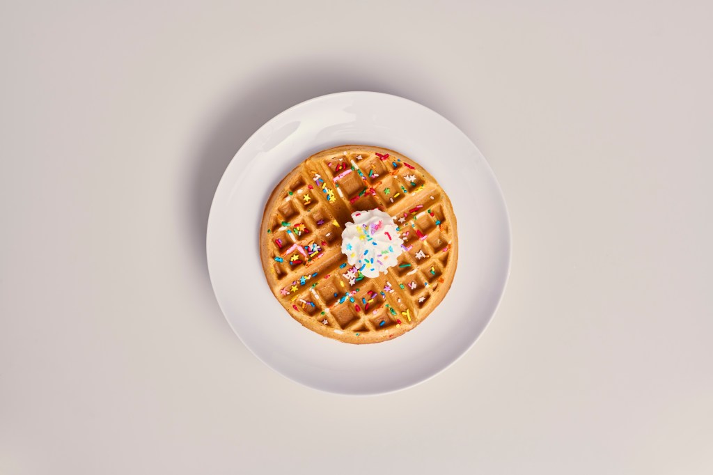 top down photo of a birthday cake flavored waffle with whipped cream and sprinkles on a white plate