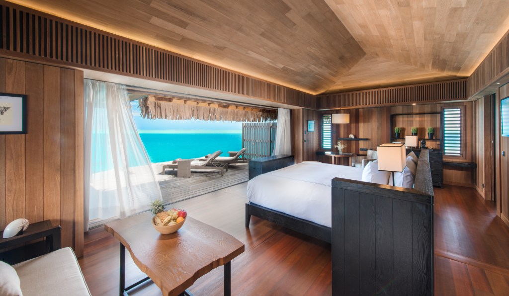 Conrad Bora Bora Nui - Over Water Villa with Pool, bed facing the ocean, couch, end table with shell, coffee table with decorative fruit bowl, shelves, lamps, deck with lounge chairs