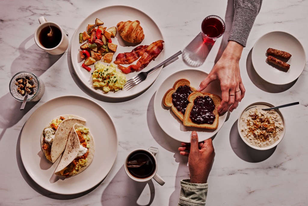 plates with Breakfast Tacos, Overnight Oats, Chia Pudding, jam on toast, sausage, tea, two people reaching for toast