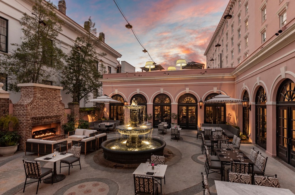 Mills House Charleston, Curio Collection by Hilton - Courtyard, dining seating area at sunset, lit up fountain in the center, outdoor brick fireplace and couches