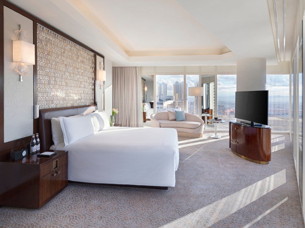Waldorf Astoria Las Vegas, bedroom with two walls of windows, TV, double bed, couch, sconces, bedside table, alarm clock, bottled water, flowers in a vase, couch end table with books