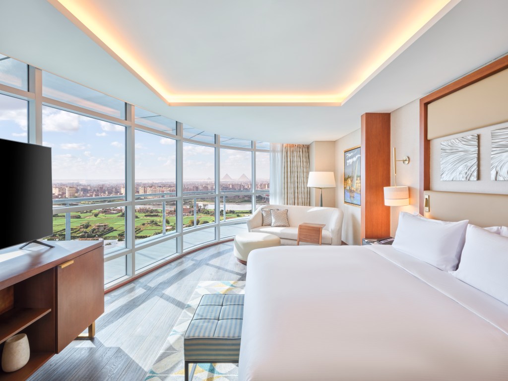 King bed, tv on a stand, white couch and ottoman, lamp and wall of windows with city and pyramid views