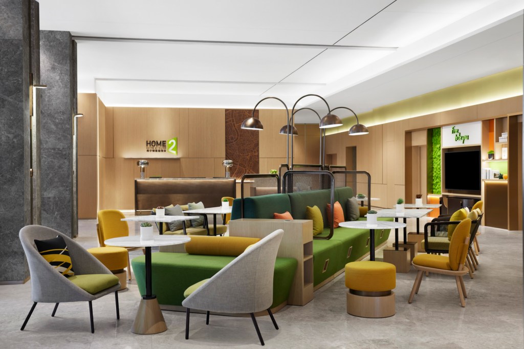 Lobby at with large seating area featuring green and yellow accents, overhead lights, tables and chairs and reception desk in Home2 Suites by Hilton Shenzhen Bao'an