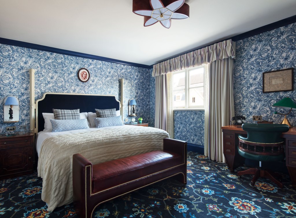 Graduate by Hilton Oxford - Guest Room, blue floral wallpaper, blue, grey, cream and white bedding, blue floral carpet, red chaise, bed side tables with small lamps with blue lampshade, vintage desk with green chair, lamp and rotary phone, flower shaped ceiling light