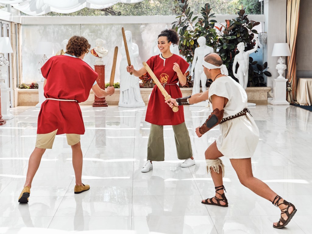 Couple in gladiator training experience with trainer, wooden swords, statuary surrounding area