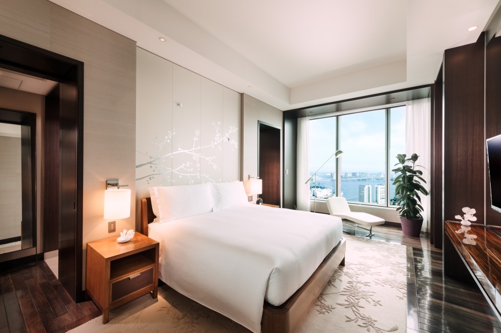 Conrad Tokyo, Deluxe Bay View Suite Bedroom, guest room, white comforter, white pillows, white chaise, wooden nightstand, potted plant, area rug, reading lamp, city view, white curtains