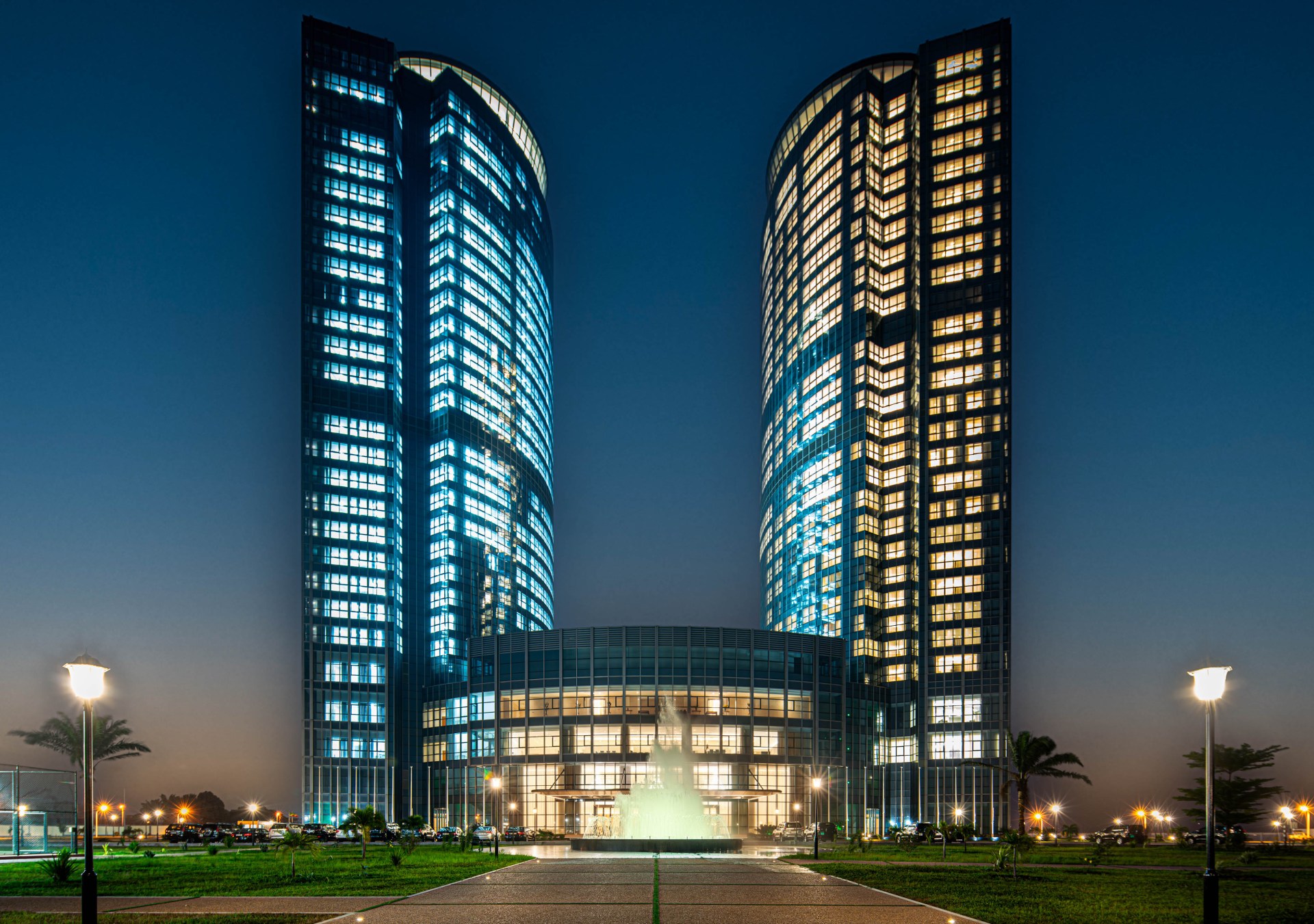 Hilton Brazzaville Les Tours Jumelles Hotel & Residences, Exterior, water fountain, sunset, palm trees, walkway, grass, parking lot, light posts