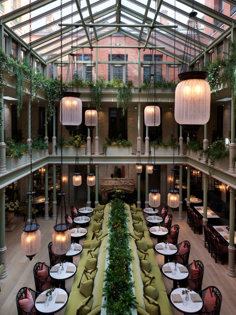 NoMad London Restaurant - view of restaurant long benches and chairs along tables in the center of the room, glass roof and hanging light fixtures, hanging vines and greenery on 2nd and third levels, fireplace at the end of the main dining hall