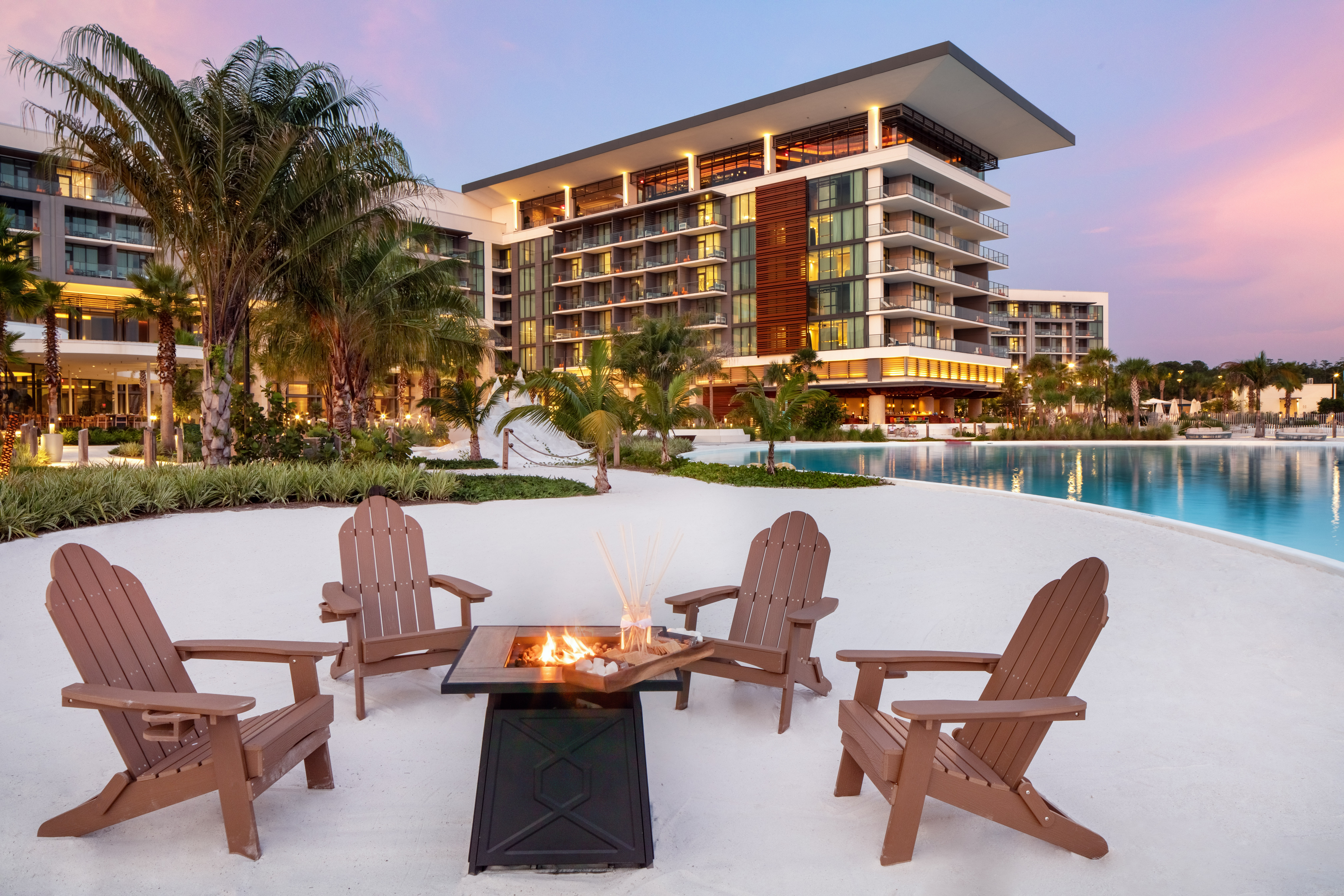 An outdoor roast fire pit surrounded by four brown adirondack chairs, white sand, a body of water, and palm trees located in front of the Conrad Orlando location