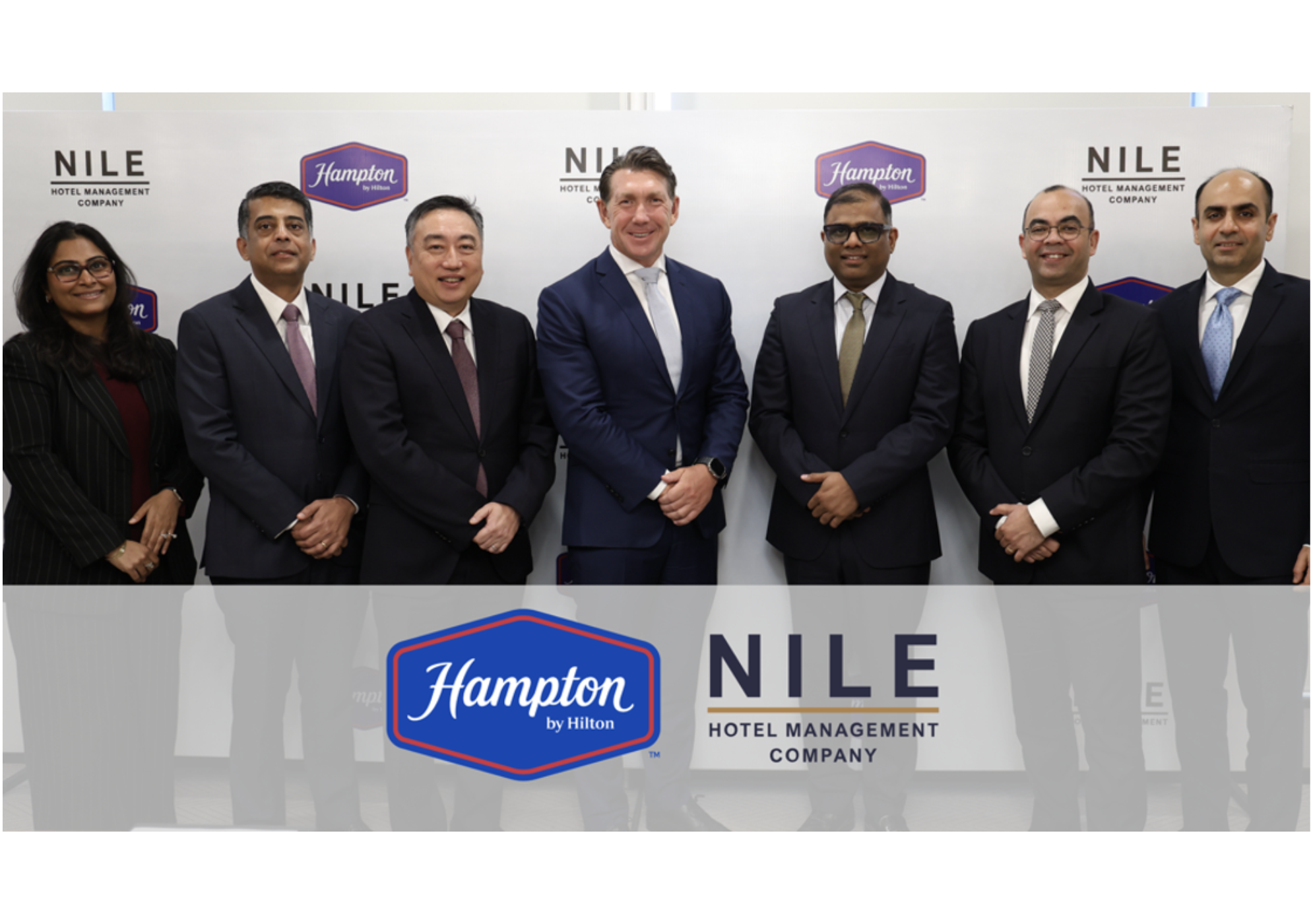 Seven people standing and smiling in front of Hampton and NILE Hotel Management Company logo backdrop at the Hampton by Hilton and NILE Hospitality Signing Ceremony