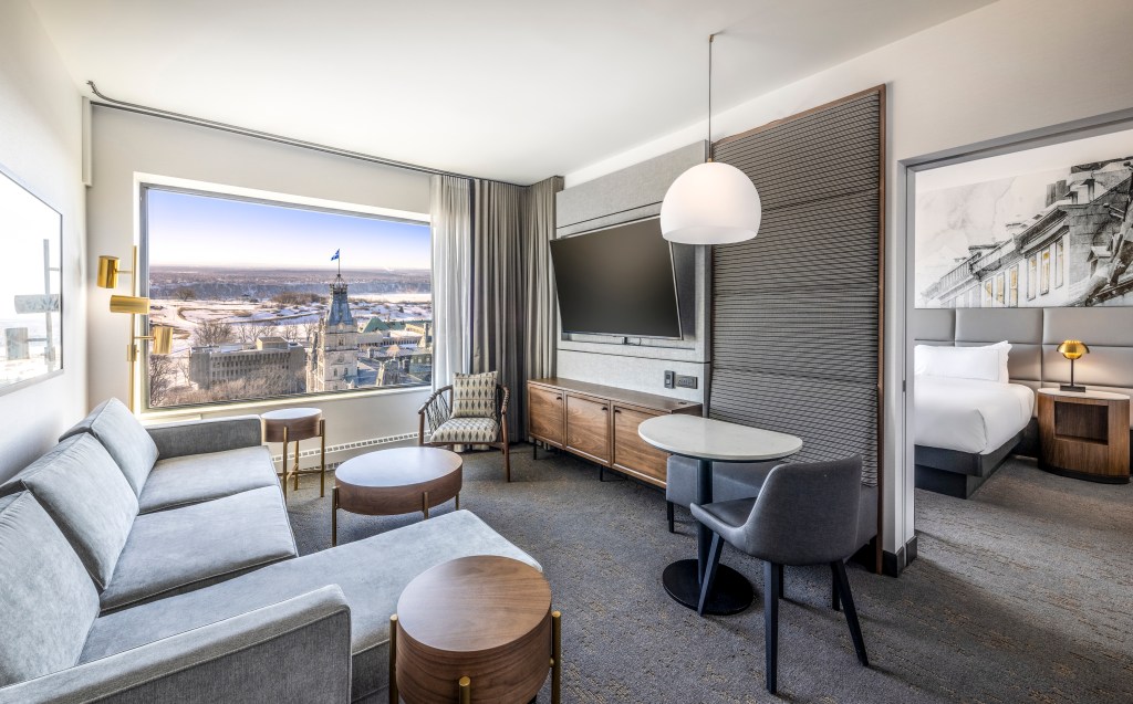 A junior suite at the Hilton Quebec location with one king bed, grey colored couch, a mounted television, big window overlooking the city, and modern lamps throughout the room