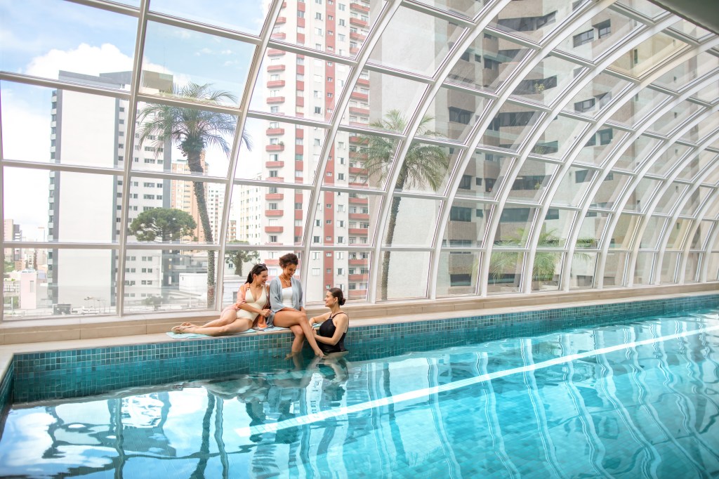 Qoya Hotel Curitiba, Curio Collection by Hilton, Indoor Pool, curved picture windows, blue tiled floor, people smiling, view of buildings, palm trees