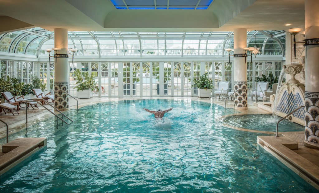 Rome Cavalieri, a Waldorf Astoria Hotel, Indoor Pool (Antonio Saba), person swimming, swimmer, curved paneled glass wall entrance, decorative tiles, water fountain, chaise lounge seating, lounge sitting area, potted plants, white planter boxes