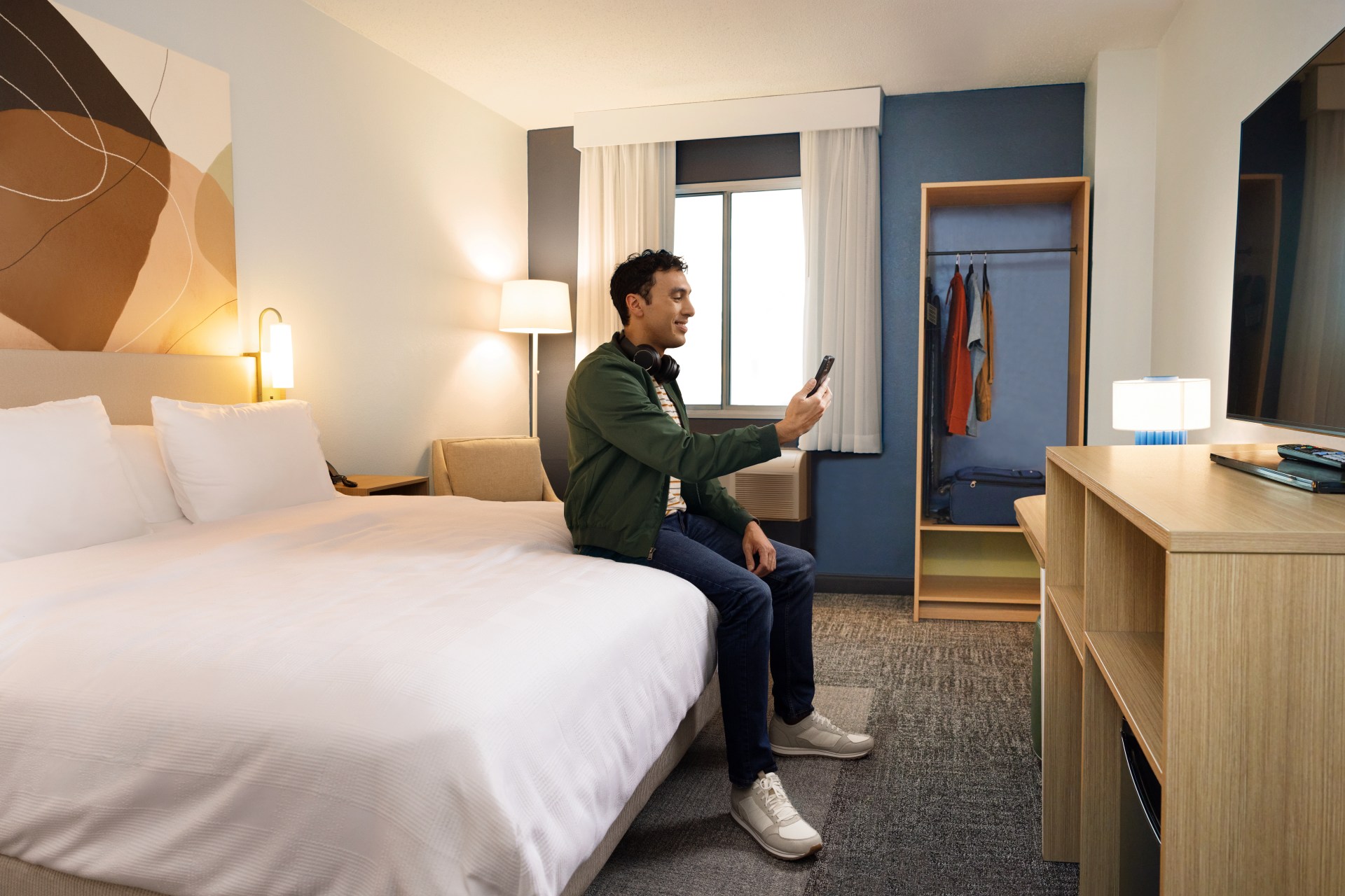 Spark by Hilton - Guest Room, guest sitting on the bed in guest room on phone, chair, clothing rack, tv and console