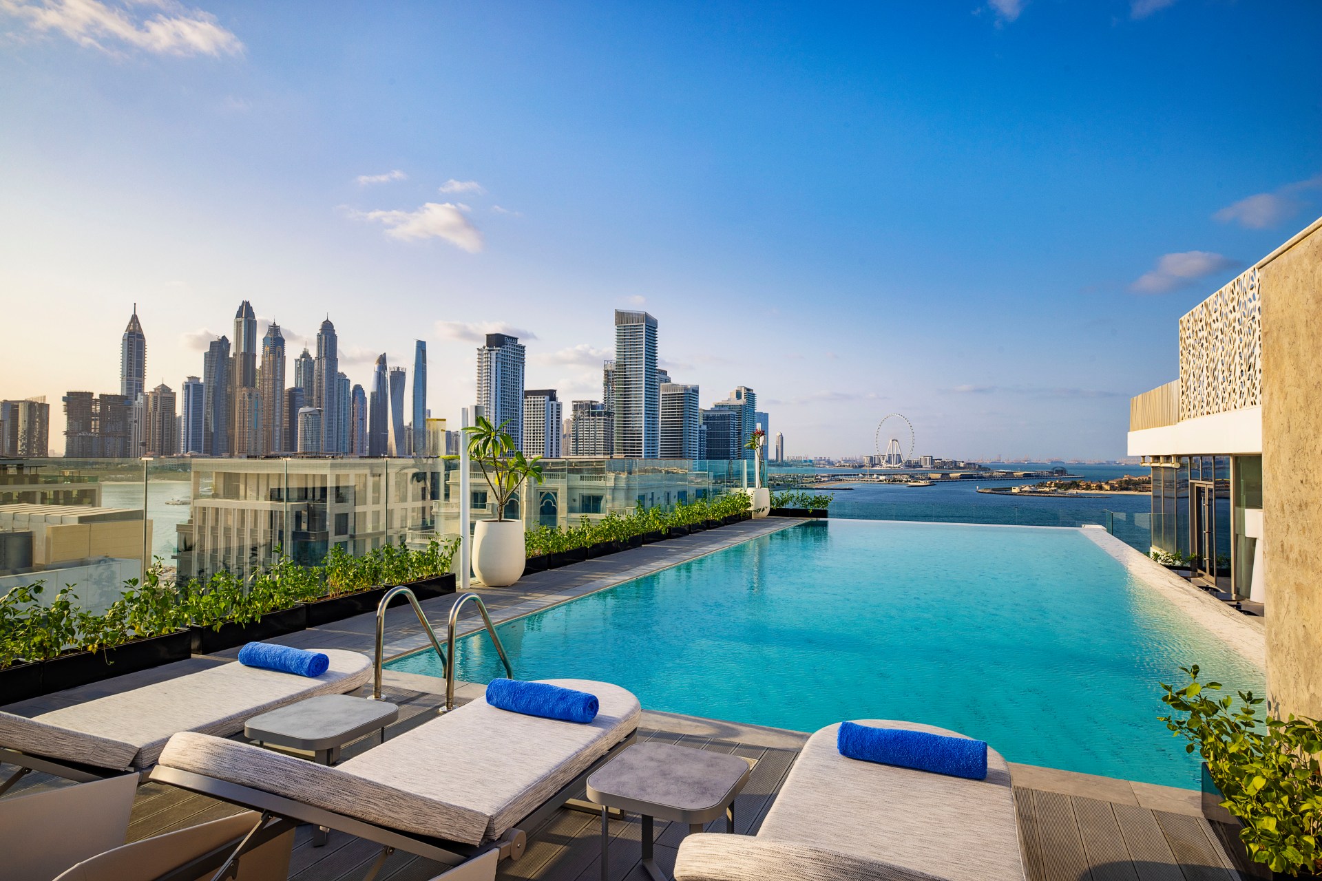 Tumbi West Palm Beach Hotel Dubai, Tapestry Collection by Hilton, Pool, grey outdoor chaise lounge chairs, royal blue towels, city view, grey end tables potted plants, infinity pool