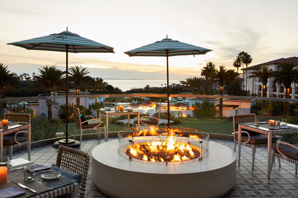 Lit outdoor fireplace at the Waldorf Astoria Monarch Beach Resort &amp; Club's AVEO Table + Bar surrounded by tables and chairs overlooking a body of water and palm trees at sunset